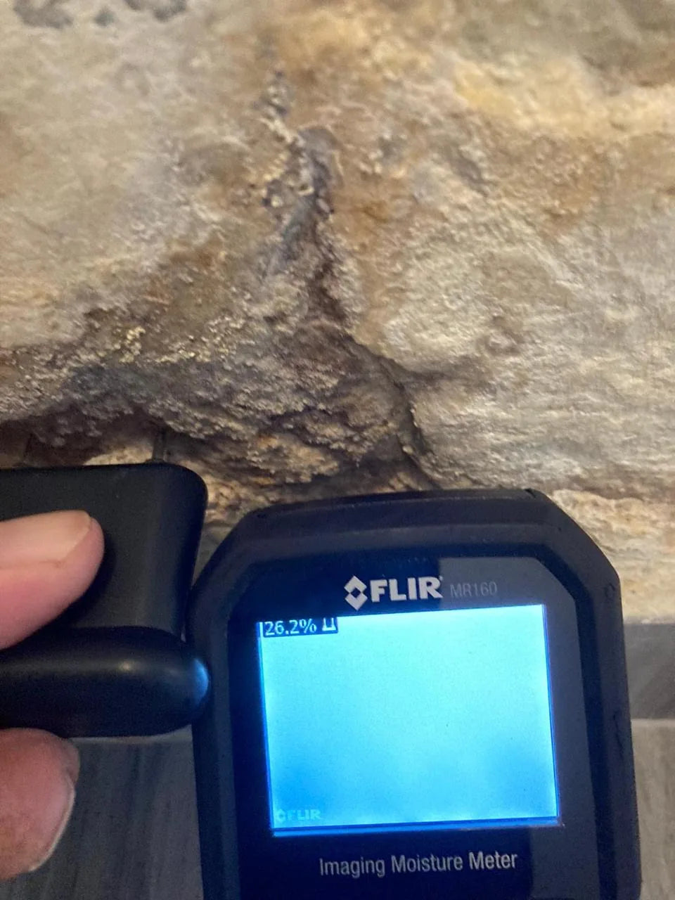 A moisture meter held against the base of a damp stone wall, reading 26.2 per cent